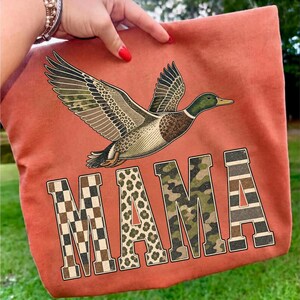 May include: A rust-colored fabric item featuring a flying mallard duck graphic above the word MAMA. The letters are filled with distinct patterns: a brown and white checkerboard, a tan leopard print, green camouflage, and horizontal stripes.