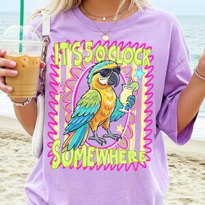 May include: A lilac-colored t-shirt featuring a colorful parrot wearing sunglasses and holding a cocktail. The shirt has the text "IT'S 5 O'CLOCK SOMEWHERE" in a playful font, surrounded by a decorative frame with stars.