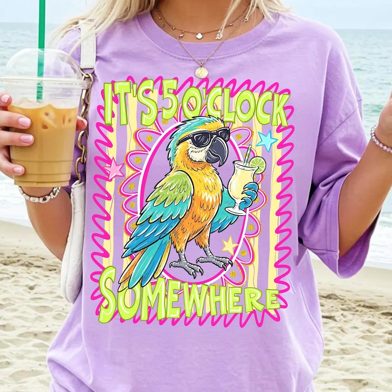 May include: A lilac-colored t-shirt featuring a colorful parrot wearing sunglasses and holding a cocktail. The shirt has the text "IT'S 5 O'CLOCK SOMEWHERE" in a playful font, surrounded by a decorative frame with stars.