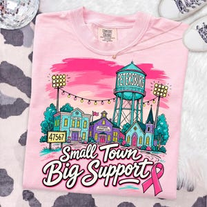 May include: A light pink t-shirt featuring a colorful graphic of a small town scene with a water tower labeled Petersburg, a city hall, a school, stadium lights, and the text Small Town Big Support with a pink awareness ribbon at the bottom.