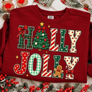Peut inclure: Un sweat-shirt &agrave; col rond rouge fonc&eacute; avec l'inscription HOLLY JOLLY en grandes lettres d&eacute;coratives. Le motif comprend un sapin de No&euml;l vert, un bonhomme en pain d'&eacute;pice, des n&oelig;uds rouges et des motifs &agrave; rayures, carreaux et pois.