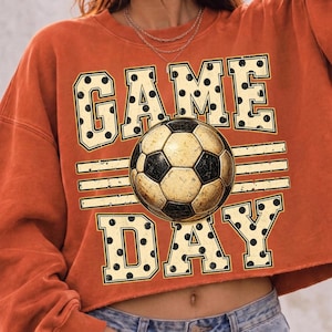 May include: A burnt orange cropped sweatshirt featuring a graphic print with the text GAME DAY in large, cream-colored block letters with black polka dots. A realistic soccer ball is centered between the words, flanked by horizontal lines.