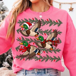 May include: A bright pink sweatshirt featuring a graphic print of three mallard ducks in flight. The ducks are decorated with a red Santa hat, a red bow, and a red and green plaid scarf, surrounded by green pine branches and red light bulbs.