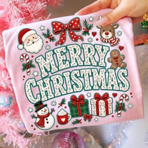 May include: A light pink t-shirt featuring the text Merry Christmas in white letters with a green outline. The design includes a Santa Claus, a reindeer, a gingerbread man, a snowman, gift boxes, candy canes, and a red bow with white dots.