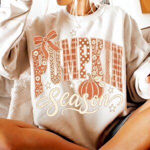 May include: A cream-colored crewneck sweatshirt featuring a large graphic print. The text PUMPKIN is written in block letters with orange plaid, floral, and polka dot patterns, accented by a bow. The word Season is written below in cursive.