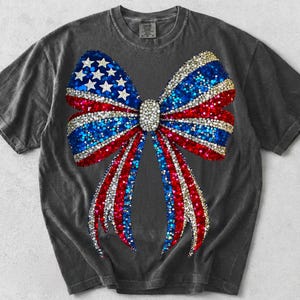 May include: A charcoal gray short-sleeve t-shirt featuring a large, central graphic of a bow. The bow is decorated with a sparkling, glitter-like pattern in red, white, and blue, incorporating white stars on a blue background to resemble a flag.