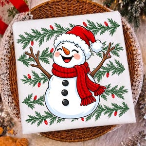 Puede incluir: Una camiseta blanca con un mu&ntilde;eco de nieve de dibujos animados con nariz de zanahoria, bufanda de punto roja y gorro de Pap&aacute; Noel rojo. El mu&ntilde;eco tiene brazos de rama y tres botones negros, rodeado de ramas de pino verdes y bombillas rojas.