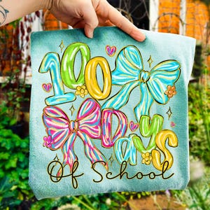 Op de afbeelding: Een turquoise vierkant met de tekst "100 Days of School" in kleurrijke, grillige letters. Het ontwerp bevat grote, decoratieve strikken in roze, blauwe en gele tinten, met bloemaccenten en harten.