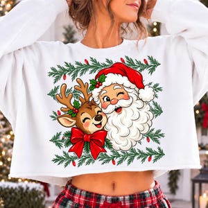 May include: A white long-sleeve cropped sweatshirt featuring a graphic print of Santa Claus and a reindeer. The design includes green pine branches, red berries, and a large red bow. Santa wears a red hat and has a white beard.