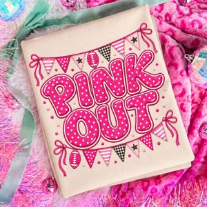 K&ouml;nnte beinhalten: Ein cremefarbenes T-Shirt mit dem Schriftzug "PINK OUT" in gro&szlig;en, kr&auml;ftigen, pinkfarbenen Buchstaben mit wei&szlig;en Punkten. Das Design wird von zwei Reihen Wimpelketten mit Fu&szlig;b&auml;llen, Sternen und Karomustern in Pink und Schwarz eingerahmt.