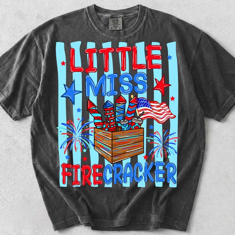 Może przedstawiać: Ciemnoszary t-shirt z kr&oacute;tkim rękawem z nadrukiem graficznym w jasnoniebieskie pionowe paski. Napis brzmi LITTLE MISS FIRECRACKER w kolorze czerwonym i niebieskim. Wz&oacute;r zawiera drewnianą skrzynkę z fajerwerkami, gwiazdkami i amerykańską flagą.