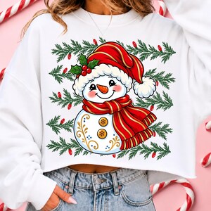 May include: A white cropped sweatshirt featuring a graphic of a snowman wearing a red Santa hat and a red scarf. The snowman has a carrot nose, two gold buttons, and decorative gold swirls. The background print includes green pine branches.
