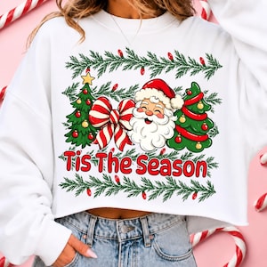 Puede incluir: Una sudadera blanca corta con un estampado gr&aacute;fico de Santa Claus, dos &aacute;rboles de Navidad decorados y un gran lazo a rayas rojas y blancas. El texto 'Tis The Season' est&aacute; impreso en rojo debajo del gr&aacute;fico, enmarcado por ramas de pino verdes.