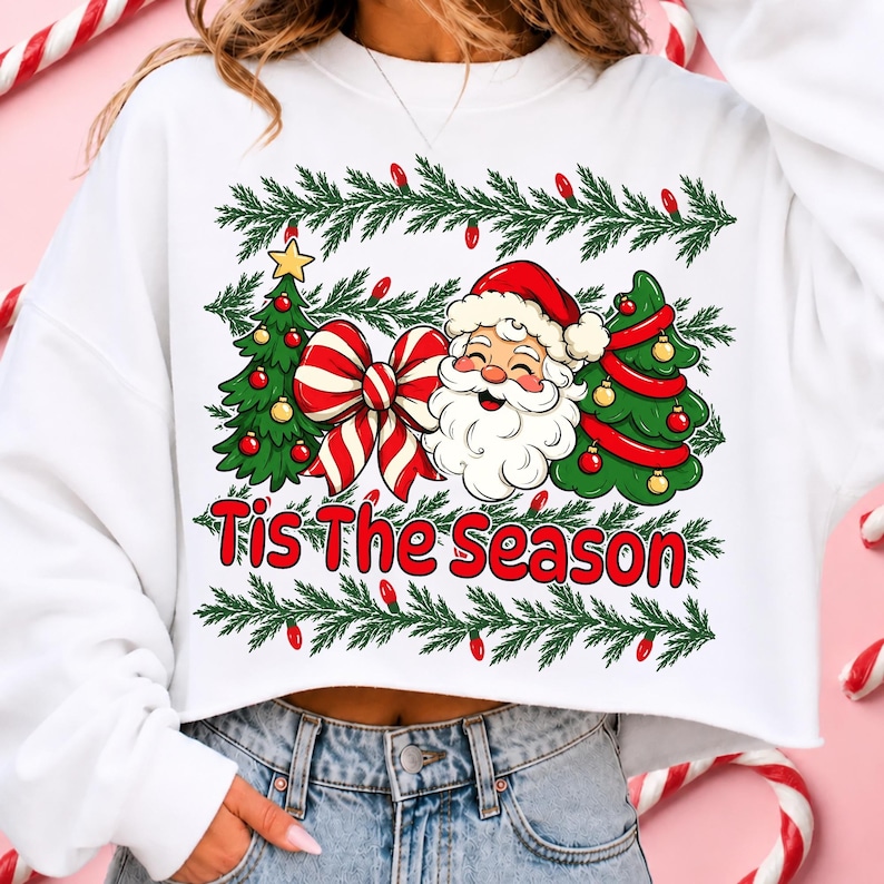 Puede incluir: Una sudadera blanca corta con un estampado gr&aacute;fico de Santa Claus, dos &aacute;rboles de Navidad decorados y un gran lazo a rayas rojas y blancas. El texto 'Tis The Season' est&aacute; impreso en rojo debajo del gr&aacute;fico, enmarcado por ramas de pino verdes.