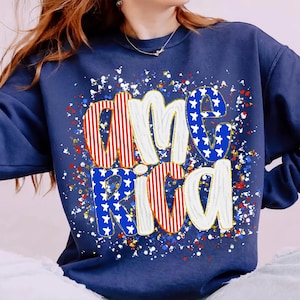 K&ouml;nnte beinhalten: Marineblaues Sweatshirt mit dem Wort "America" in einem patriotischen Design. Die Buchstaben sind wei&szlig; mit roten und blauen Akzenten, einschlie&szlig;lich Sternen und Streifen. Das Design ist von roten, wei&szlig;en und blauen Sprenkeln umgeben.