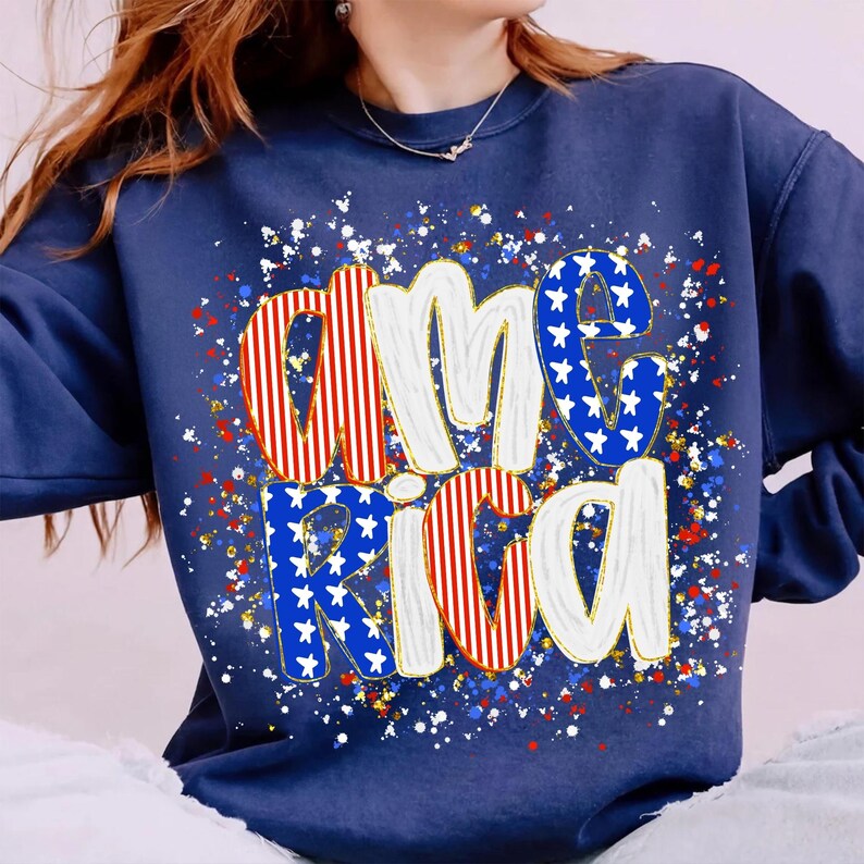 K&ouml;nnte beinhalten: Marineblaues Sweatshirt mit dem Wort "America" in einem patriotischen Design. Die Buchstaben sind wei&szlig; mit roten und blauen Akzenten, einschlie&szlig;lich Sternen und Streifen. Das Design ist von roten, wei&szlig;en und blauen Sprenkeln umgeben.