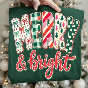 May include: A dark green t-shirt featuring the text MERRY in large block letters with varied patterns including red bows, green plaid, red and white stripes, pink and green trees, and white polka dots, with the red script text & bright below.
