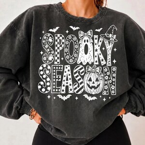 May include: A charcoal grey crewneck sweatshirt featuring a white graphic print that reads SPOOKY SEASON. The text is decorated with patterns including polka dots, checkers, stripes, flowers, stars, a ghost with a bow, and a jack-o'-lantern.