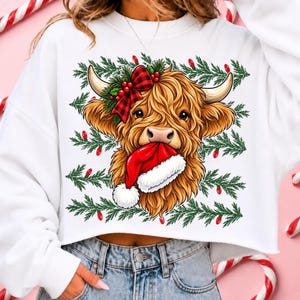 May include: A white long-sleeve sweatshirt featuring a graphic of a brown Highland cow with a red and black plaid bow, green pine branches, and red berries. The cow is holding a red and white Santa hat in its mouth against a pine background.