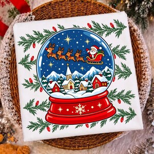 May include: A white fabric square featuring a graphic of a snow globe with a red base. Inside the globe, Santa Claus in a sleigh pulled by reindeer flies over a snowy village with houses and a church. Green pine branches with red lights frame it.