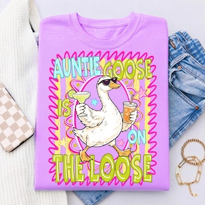 K&ouml;nnte beinhalten: Ein helllila T-Shirt mit dem Text "Auntie Goose is on the Loose". Die Grafik zeigt eine Comic-Gans mit Sonnenbrille, die eine Margarita und ein Getr&auml;nk mit Strohhalm h&auml;lt. Das Design ist von einem farbenfrohen Rahmen umgeben.