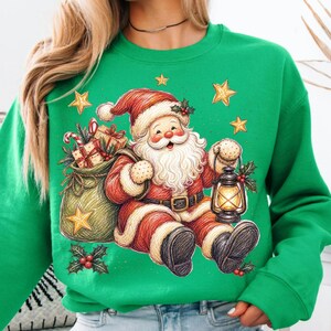 K&ouml;nnte beinhalten: Ein leuchtend gr&uuml;ner Rundhals-Pullover mit einer grafischen Illustration des Weihnachtsmanns in einem roten Anzug mit wei&szlig;em Pelzbesatz, der eine leuchtende Laterne h&auml;lt. Daneben steht ein gr&uuml;ner Sack voller verpackter Geschenke und Zuckerstangen.