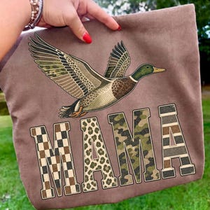 May include: A brown t-shirt featuring a graphic of a flying mallard duck with camouflage and leopard print wings. Below the duck, the word MAMA is printed in large block letters, with each letter featuring a different pattern: checkerboard, leopard, camouflage, and horizontal stripes.