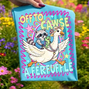 May include: A light blue t-shirt featuring a whimsical illustration. A raccoon wearing overalls and glasses rides on a goose with a frog passenger. The text reads "OFF TO CAWSE A FERFUFFLE" in a playful font.