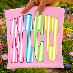 以下が含まれることがあります： 大きな太字のアーチ状フォントで「NICU」という文字が描かれたライトピンクの布製ポーチ。各文字は垂直の白いストライプ模様で塗りつぶされており、Nはピンク、Iは青、Cは緑、Uは黄色という異なる色で縁取られています。