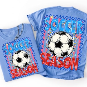 May include: A light blue t-shirt featuring a graphic print of a black and white football centered above the red text "SEASON" and below the blue text "SOCCER". The design includes pink vertical stripes, blue squiggly borders, and small stars.