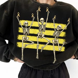 May include: A black crewneck sweatshirt featuring a graphic print of three dancing skeletons. Each skeleton is holding a small yellow ribbon above its head. The skeletons are positioned in front of three horizontal, distressed yellow brush strokes.