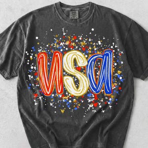 Może przedstawiać: Ciemnoszary t-shirt z kr&oacute;tkim rękawem z nadrukiem graficznym liter USA w stylizowanej, zaokrąglonej czcionce. Litery są czerwone, białe i niebieskie, otoczone żywym wzorem rozprysk&oacute;w czerwonej, białej, niebieskiej i złotej farby.