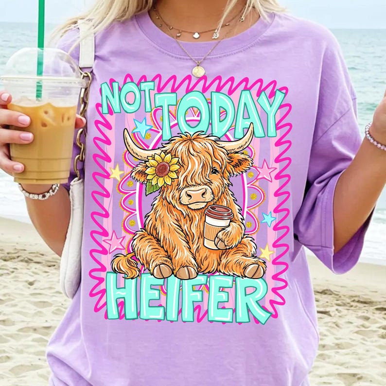May include: A lilac-colored t-shirt features a graphic of a Highland cow with a sunflower and a coffee cup. The text reads "NOT TODAY HEIFER" in pink and turquoise. The design includes stars and wavy lines.