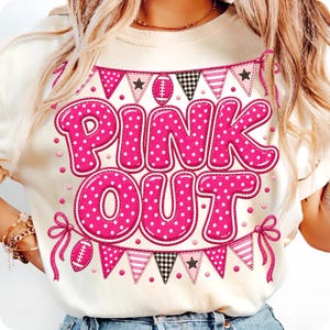 K&ouml;nnte beinhalten: Ein cremefarbenes T-Shirt mit einem Grafikdruck, der den Text PINK OUT in gro&szlig;en, rosa, wei&szlig; gepunkteten Buchstaben zeigt. Das Design umfasst rosa Wimpelketten mit Sternen, Streifen und Footballs, die an den Enden mit rosa Schleifen gebunden sind.