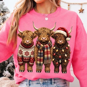 May include: A bright pink crewneck sweatshirt featuring a graphic of three brown Highland cattle. The left cow wears a red and white snowflake sweater, the center cow wears a plaid scarf, and the right cow wears a Santa hat and Christmas lights.