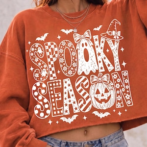 May include: An orange long-sleeve sweatshirt featuring a white graphic print that reads SPOOKY SEASON. The text is decorated with patterns including flowers, checkers, and stripes, alongside illustrations of a ghost, a pumpkin, and bats.