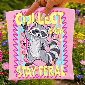 Op de afbeelding: Een lichtroze stoffen item met een afbeelding van een grijze wasbeer die een rock-and-roll handgebaar maakt. De tekst luidt COLLECT DATA STAY FERAL in gele letters. Het ontwerp bevat roze kronkelige randen, blauwe strepen en sterren.