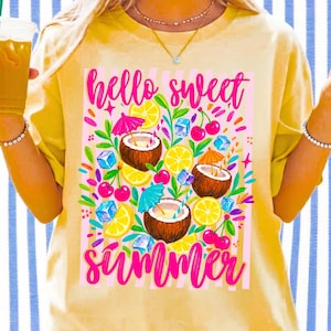 May include: A pale yellow t-shirt featuring a graphic print with the text "hello sweet summer" in pink script. The design includes illustrations of three coconuts with colorful cocktail umbrellas, lemon slices, cherries, and blue ice cubes.