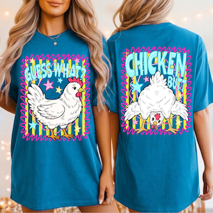 Op de afbeelding: Een blauwgroen t-shirt met een grafische print van een witte kip op de voorkant met de tekst "GUESS WHAT?" en een voorovergebogen witte kip op de achterkant met de tekst "CHICKEN BUTT". Het ontwerp bevat gele verticale strepen en sterren.