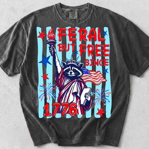Może przedstawiać: Ciemnoszary t-shirt z kr&oacute;tkim rękawem z grafiką szopa pracza przebranego za Statuę Wolności. Napis głosi FERAL BUT FREE SINCE 1776. Projekt zawiera pochodnię, książkę, amerykańską flagę, niebieskie paski oraz czerwone gwiazdy.