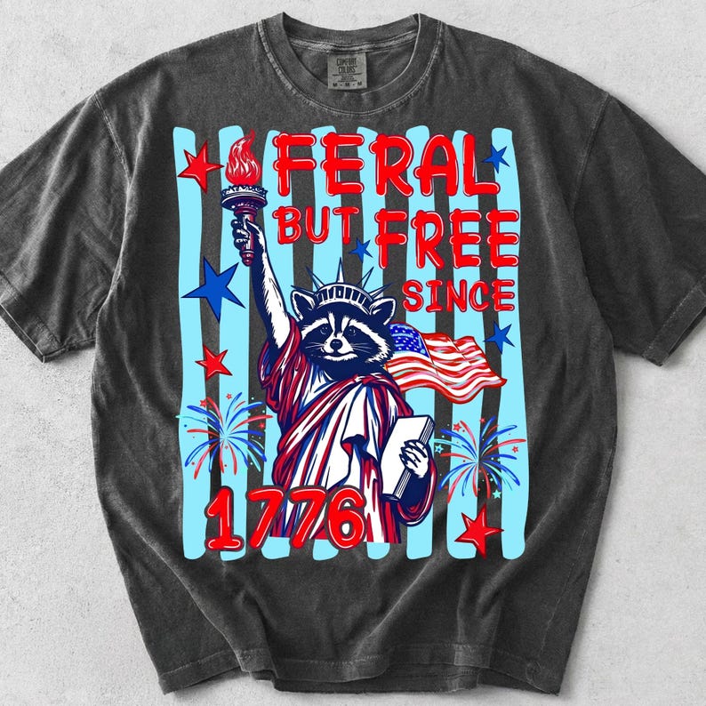 Może przedstawiać: Ciemnoszary t-shirt z kr&oacute;tkim rękawem z grafiką szopa pracza przebranego za Statuę Wolności. Napis głosi FERAL BUT FREE SINCE 1776. Projekt zawiera pochodnię, książkę, amerykańską flagę, niebieskie paski oraz czerwone gwiazdy.