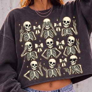 May include: A charcoal gray crewneck sweatshirt featuring a graphic print of nine skeletons in various poses, interspersed with small white bows and four-pointed stars. The skeletons are depicted with different hand gestures, including peace signs and pointing.