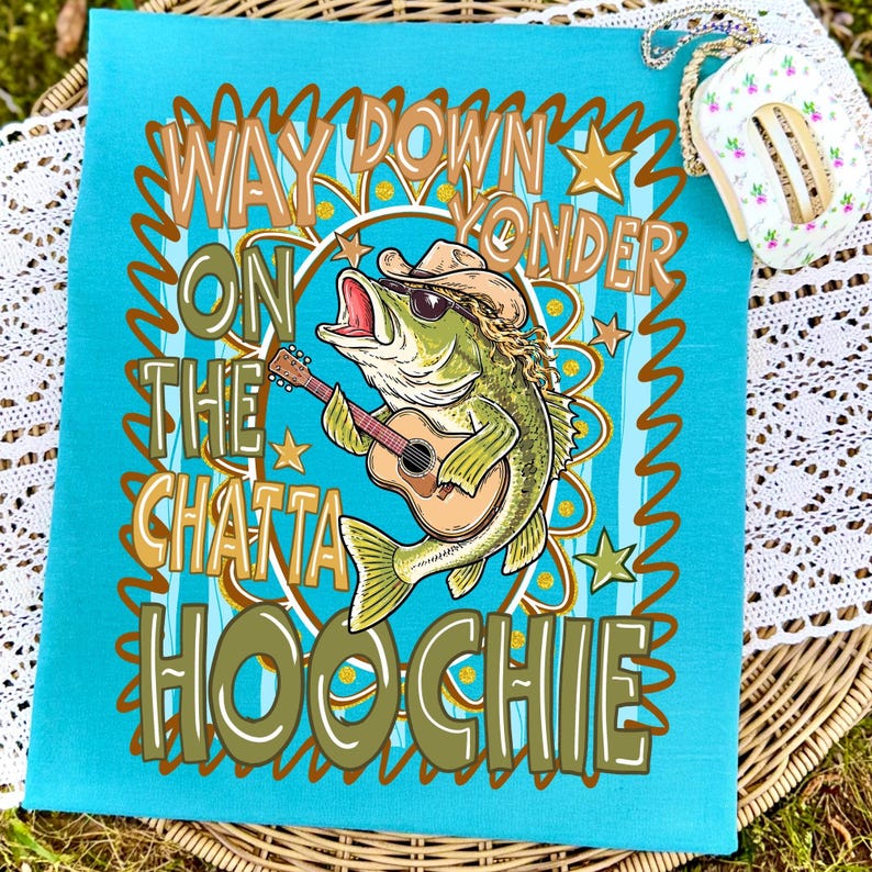 May include: A turquoise blue towel with a graphic of a fish wearing a cowboy hat and sunglasses, playing a guitar. The text on the towel reads "Way Down Yonder On The Chatta Hoochie."