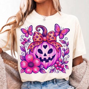 K&ouml;nnte beinhalten: Ein cremefarbenes T-Shirt mit einem Grafikdruck eines leuchtend pinken K&uuml;rbisgesichts mit herzf&ouml;rmiger Nase, das mit einer Schleife im Leopardenmuster verziert ist. Das Design umfasst zwei pinke Schmetterlinge, eine gro&szlig;e Blume und Ranken.