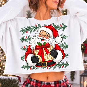 May include: A white cropped sweatshirt featuring a graphic print of Santa Claus with a white beard and red hat, holding a gold gift box with a red bow. The design is framed by green pine branches with small red lights, set against a white fabric.