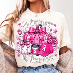 May include: A cream-colored t-shirt featuring a graphic print with the text In October We Wear Pink. The design includes a large pink pumpkin with a bow, a smaller white polka-dot pumpkin, a pink coffee cup, a pink baseball cap, and roses.