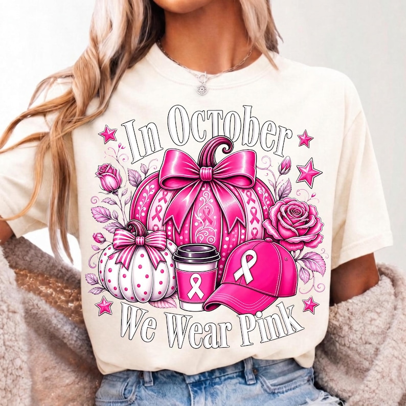 May include: A cream-colored t-shirt featuring a graphic print with the text In October We Wear Pink. The design includes a large pink pumpkin with a bow, a smaller white polka-dot pumpkin, a pink coffee cup, a pink baseball cap, and roses.