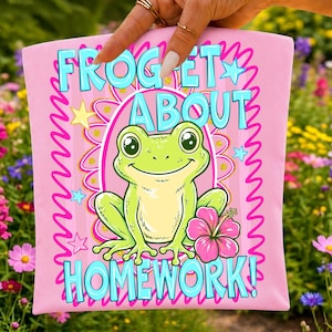 Pu&ograve; includere: Un articolo in tessuto rosa chiaro con un'illustrazione centrale di una rana verde, un fiore di ibisco rosa e il testo FROGET ABOUT HOMEWORK in lettere blu brillante. Il design include bordi ondulati rosa e piccole stelle gialle e blu.