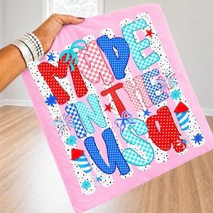 May include: A pink fabric square featuring the text MADE IN THE USA in large, multi-coloured, patterned letters. The design includes red, white, and blue polka dots, checkered patterns, and small firework and flower graphics on a white dotted background.