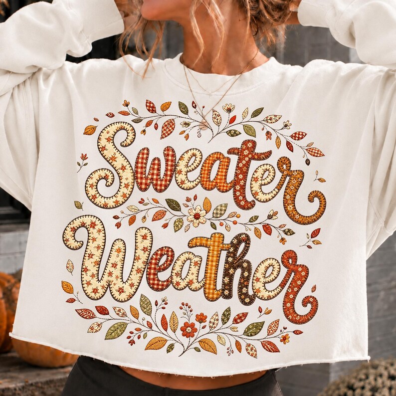 Pu&ograve; includere: Una felpa bianca a maniche lunghe e taglio corto con la scritta "Sweater Weather" in grandi lettere decorative a motivo scozzese. Il design &egrave; circondato da una disposizione circolare di foglie autunnali colorate, viticci e piccoli fiori.
